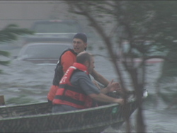 Two men wearing orange life vests navigate a small boat through flooded waters near parked cars.