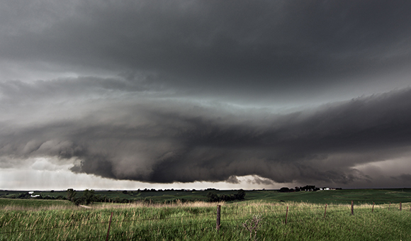 Dark, heavy storm clouds loom over a vast green landscape and grassy field under a gray sky.