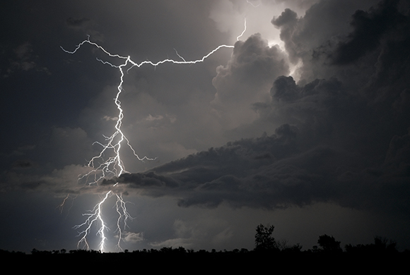 A bright lightning bolt branches across a dark, cloudy night sky over a silhouetted horizon.