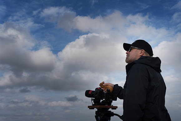 A man in a black jacket and cap looks up at a blue sky filled with large white and grey clouds.