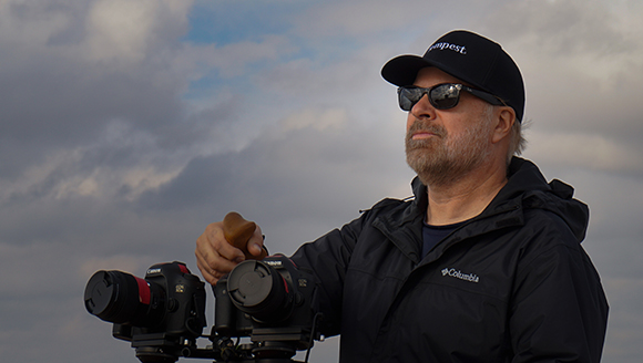 A man wearing a black cap, sunglasses, and a Columbia jacket looks toward the sky.