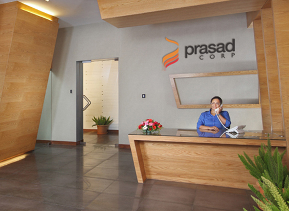A woman in a blue shirt sits at a wooden reception desk, talking on a telephone in a modern office lobby.