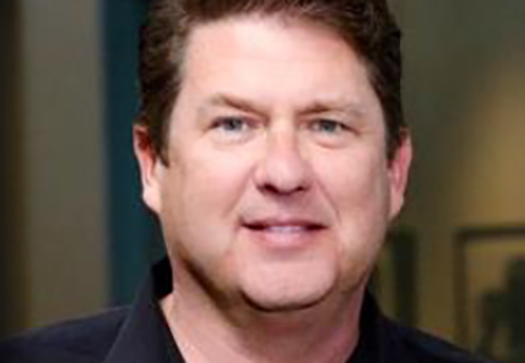 A man with short brown hair and blue eyes smiling against a blurred background.