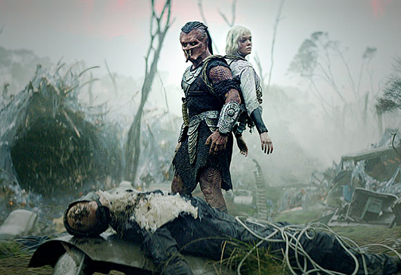 A muscular alien and a blonde woman stand over a body in a foggy, debris-filled landscape.