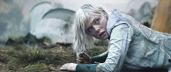 A blonde woman in a light-colored jacket crawls through dirt and grass in a hazy landscape.