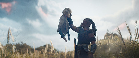 A person in dark armor holds a small blonde child up in the air in a grassy field.