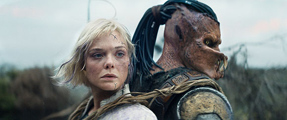 A blonde woman with facial scars looks forward next to a large, brown-skinned creature with dreadlocks.