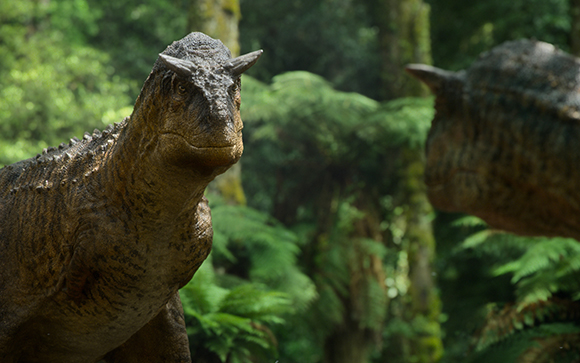 A close-up of a brown dinosaur with small horns on its head in a dense, green forest.