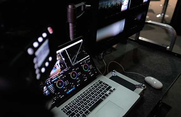 A laptop running video editing software sits on a desk next to large professional video monitors.