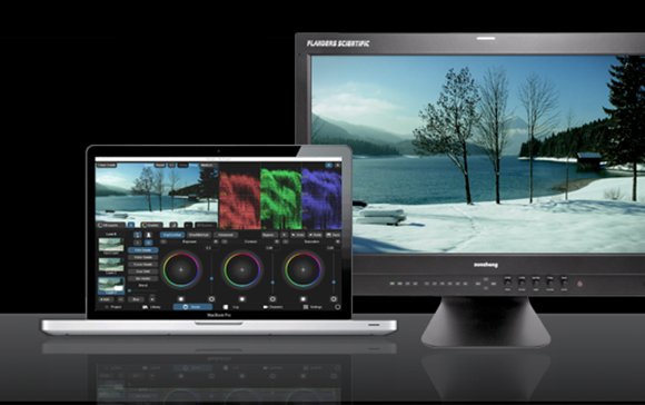 A laptop showing color grading software next to a large monitor displaying a snowy lake landscape.