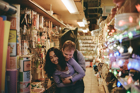 A man hugs a smiling woman from behind while standing in a narrow aisle of a well-stocked retail store.
