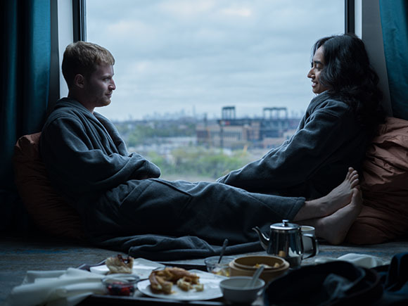 A man and woman in grey robes sit on a window ledge facing each other with a city view behind them.