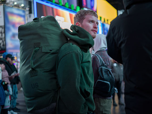 A man in a green hoodie carries a large green backpack in a busy urban setting with bright lights.