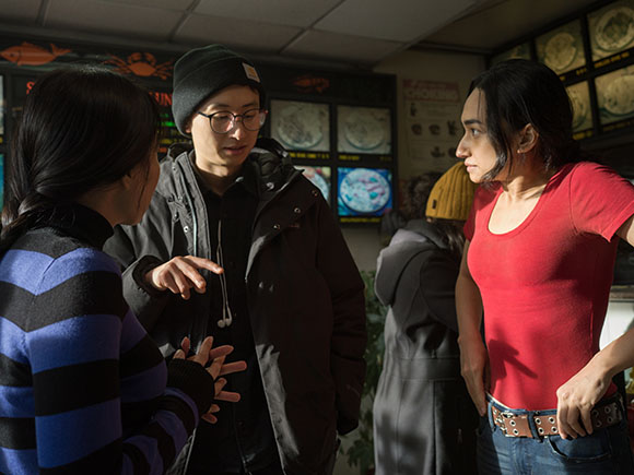 A person in a black jacket talks to a person in a red shirt near a wall with monitors.