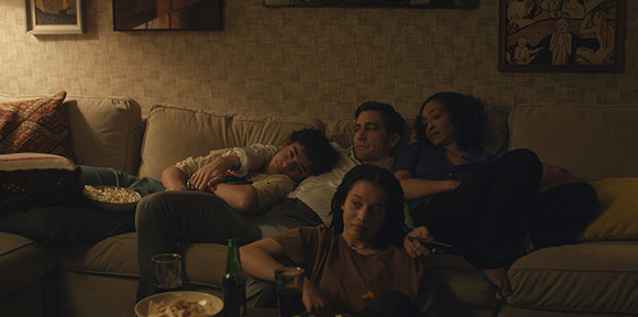 Four people lounge on a large sofa in a dark room with snacks and drinks nearby.
