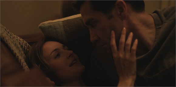 A man gazes down at a woman lying in bed as he touches her face.