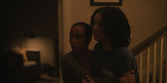 Two women stand close together in a dimly lit indoor setting near a staircase.
