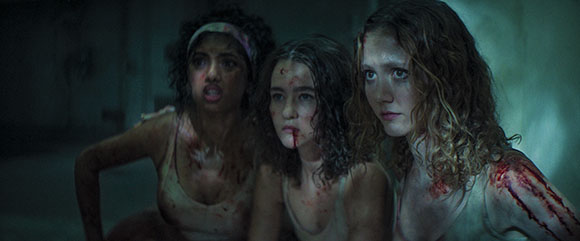 Three young women with blood on their faces and bodies look ahead with expressions of fear and distress.