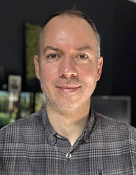 A man with short hair and light stubble wearing a grey plaid button-down shirt.