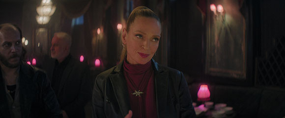 A woman with blonde hair pulled back smiles slightly in a dark, moody room with glowing pink lights.