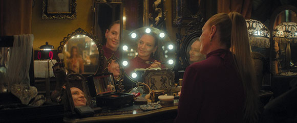 A woman in a maroon top sits at a cluttered vanity with several decorative mirrors and lights.