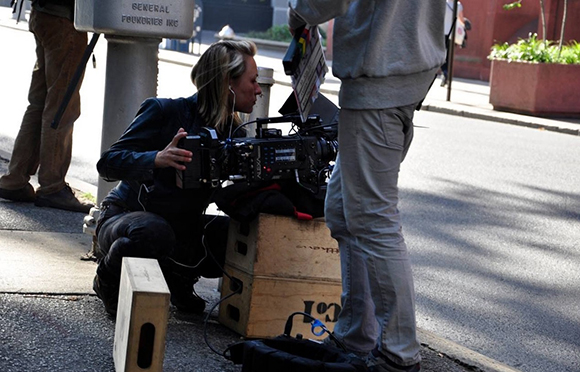 A woman crouches behind a large professional video camera mounted on a wooden crate on a city sidewalk.