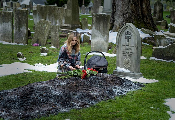 A woman in a patterned jacket places red roses on a fresh grave in a cemetery with patches of snow.