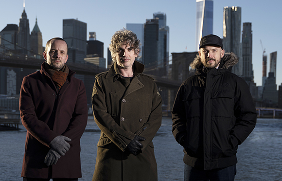 Three men in winter coats standing outdoors with a city skyline and bridge in the background.