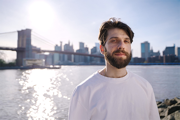 A man with a beard wearing a white t-shirt, with the Brooklyn Bridge and city skyline in the background.