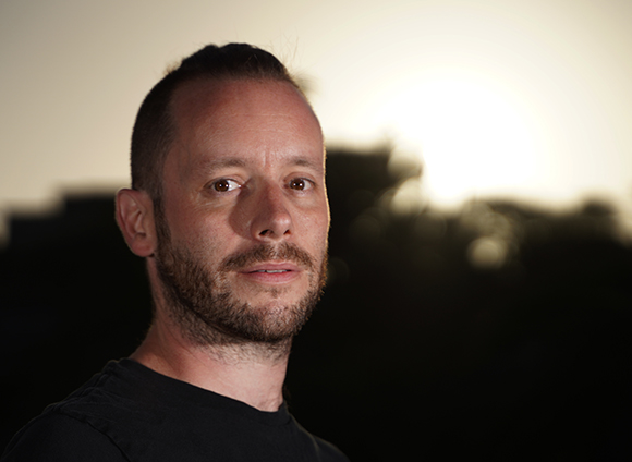 Close-up portrait of a man with short hair and a beard, looking forward against a bright sunset.