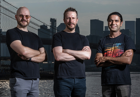 Three men standing with arms crossed in front of a city skyline and a large bridge over water.