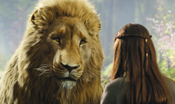 A large male lion with a thick mane looks forward next to a woman with long brown hair seen from behind.