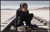 A person in black clothing crouches on railroad tracks in a vast, sandy desert under a clear sky.