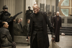 A man in dark, layered clothing stands in a large hall with armored guards and people in the background.