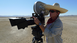 A person in a wide-brimmed hat and sunglasses operates a large professional video camera in a desert.