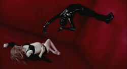 Two figures, one in a black bodysuit and one in black lingerie, lie on the floor of a deep red room.