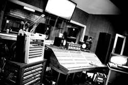 A black and white wide shot of a music recording studio featuring a large mixing console and audio equipment.
