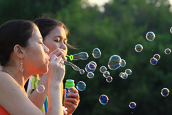 Two young girls blowing soap bubbles through plastic wands in a grassy outdoor setting.
