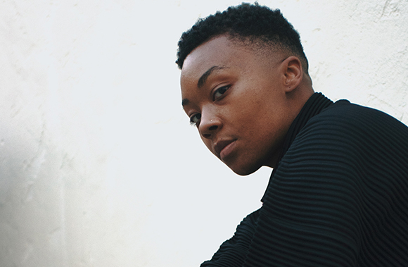 A person with short dark hair wearing a black ribbed turtleneck looking toward the camera.
