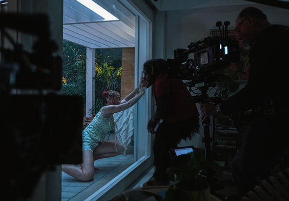 A person in a dark primate costume reaches toward a woman kneeling outside a glass door.