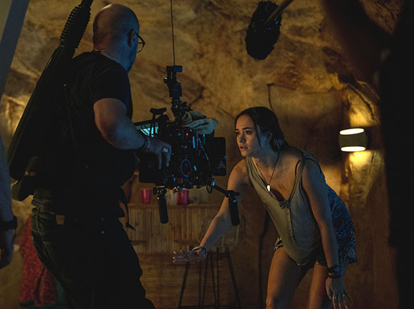 A camera operator films a woman crouching in a dimly lit cave setting.