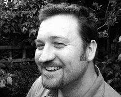 Black and white close-up portrait of a smiling man with short hair and a goatee against a leafy background.