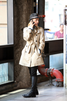 A woman in a tan trench coat and black boots talks on a cell phone while standing near a window.