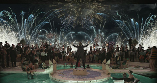 A person stands on a circular platform before a large crowd under a sky filled with fireworks.