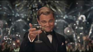 A man in a black tuxedo smiles and holds up a champagne coupe in a toast against a festive background.