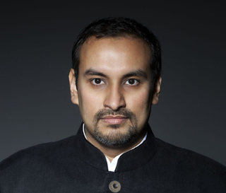 A man with dark hair and a goatee wearing a black high-collared jacket against a dark background.