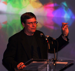 A man with glasses and a black jacket speaking into microphones at a podium.
