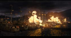 Several large wooden sailing ships burning intensely in dark waters under a cloudy night sky.
