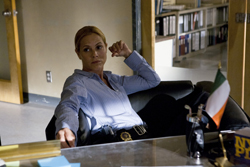 A blonde woman in a light blue shirt sits in a black office chair behind a desk in an office.