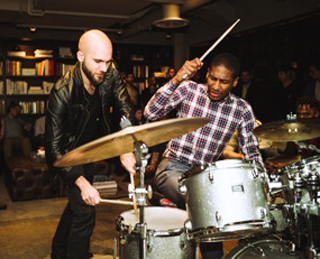 A man in a black leather jacket and a man in a plaid shirt play drums together in a dark room.